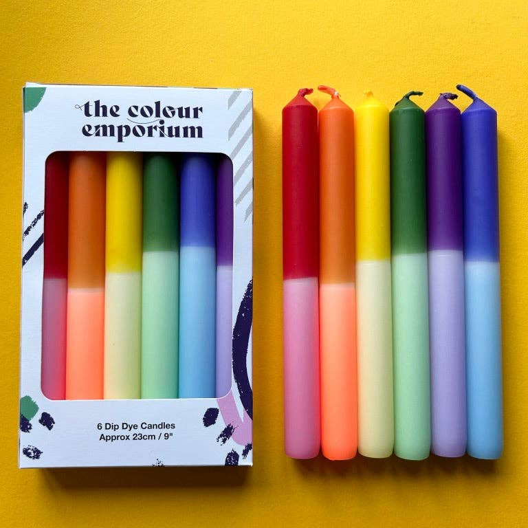 Six double-dipped dye candles in a rainbow of colors, shown next to their retail packaging box on a bright yellow background. The box is labeled 'the colour emporium'.