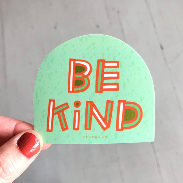 Green sticker with 'BE KIND' text held by a hand on a gray background