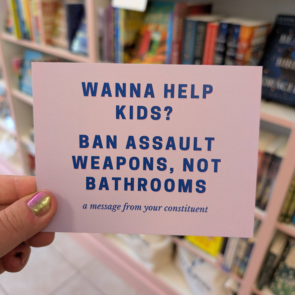 Card with text 'Wanna help kids? Ban assault weapons, not bathrooms' held in front of a bookshelf.