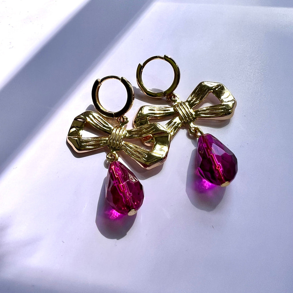 Gold bow-shaped earrings with pink teardrop charms on a light background