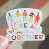 Sticker with colorful people illustration and text 'We're all in this together' held by a hand.